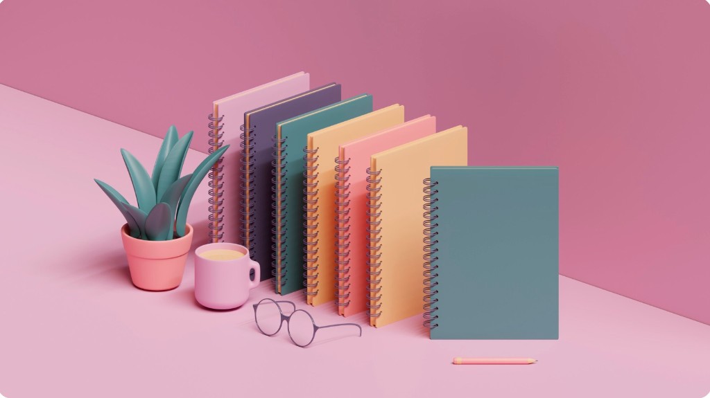 Colorful spiral-bound workbooks arranged on a pink desk with plant, mug, and glasses